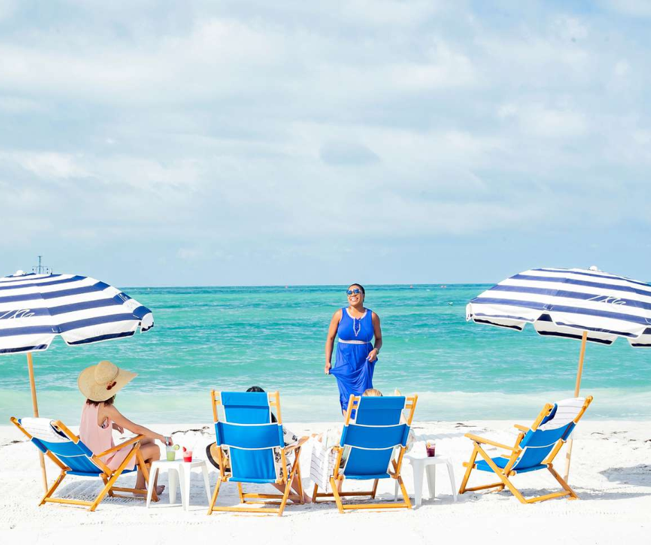 Group of people relaxing on white sand beach under striped umbrellas and blue lounge chairs by the turquoise ocean.